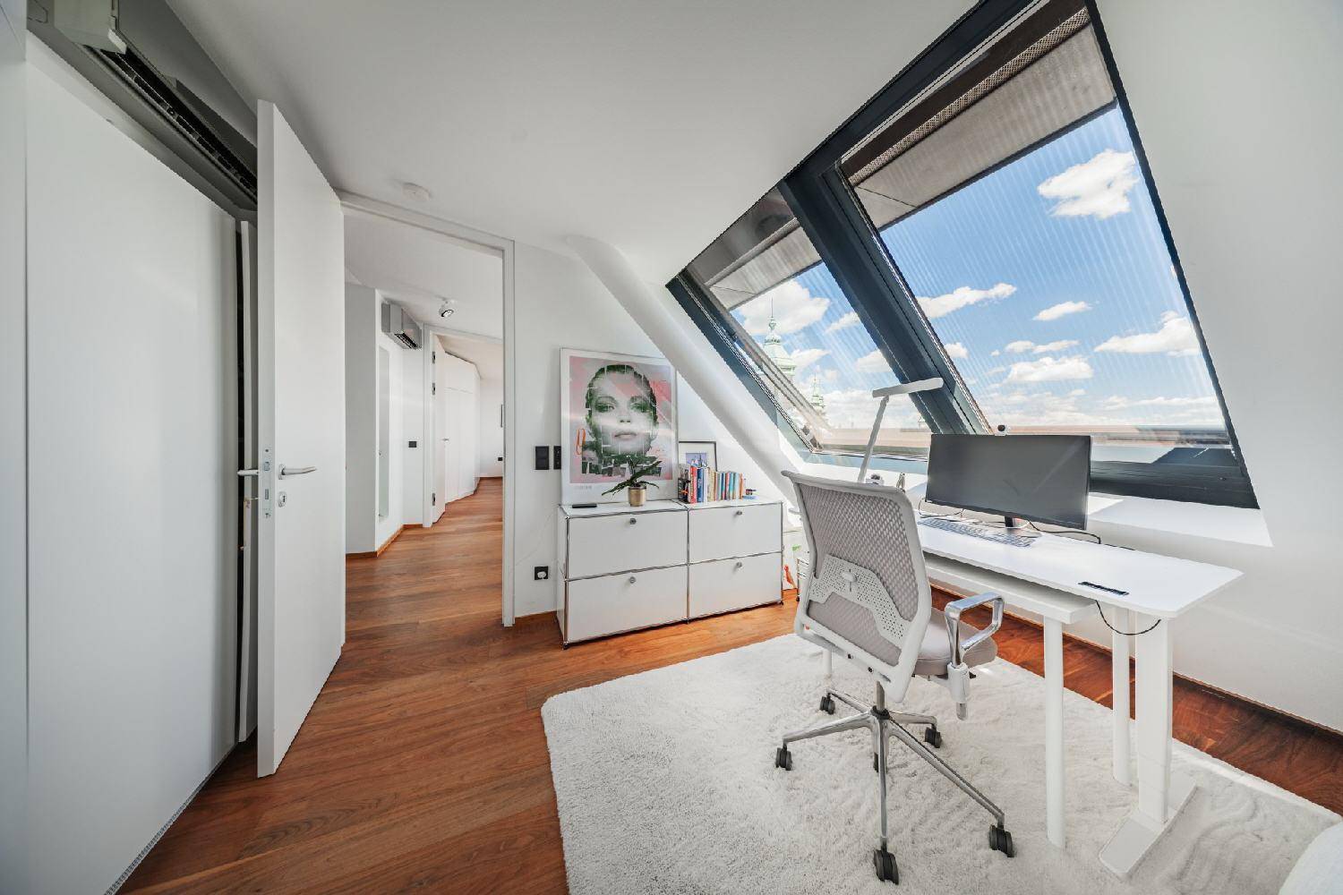 Helles Arbeitszimmer mit Holzboden, Schreibtisch und schrägen Fenstern mit Stadtblick.