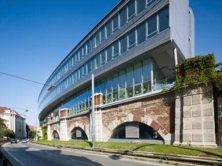 Geschwungenes Bürogebäude mit Glasfront und darunterliegender historischer Bogenkonstruktion an einer Straße.