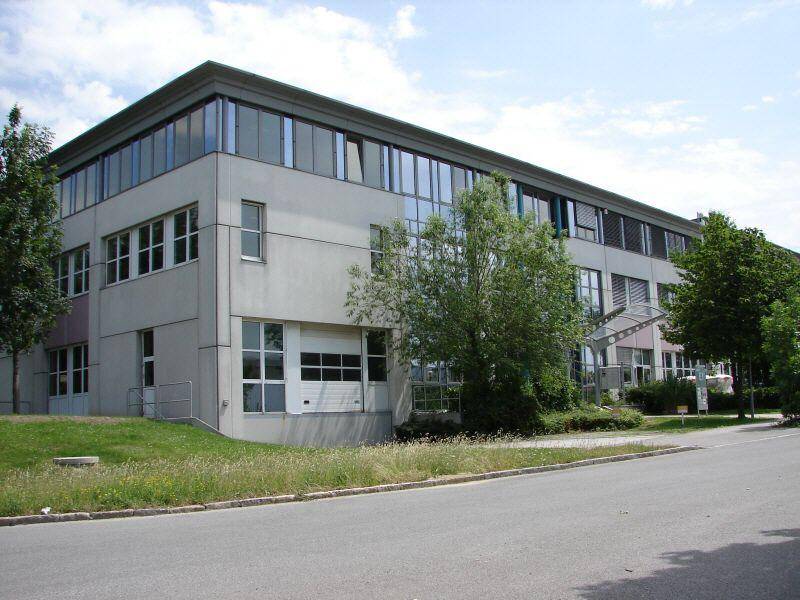 Modernes Geschäftsgebäude mit großen Fensterfronten und einer Zufahrt, umgeben von Bäumen und Rasenflächen.