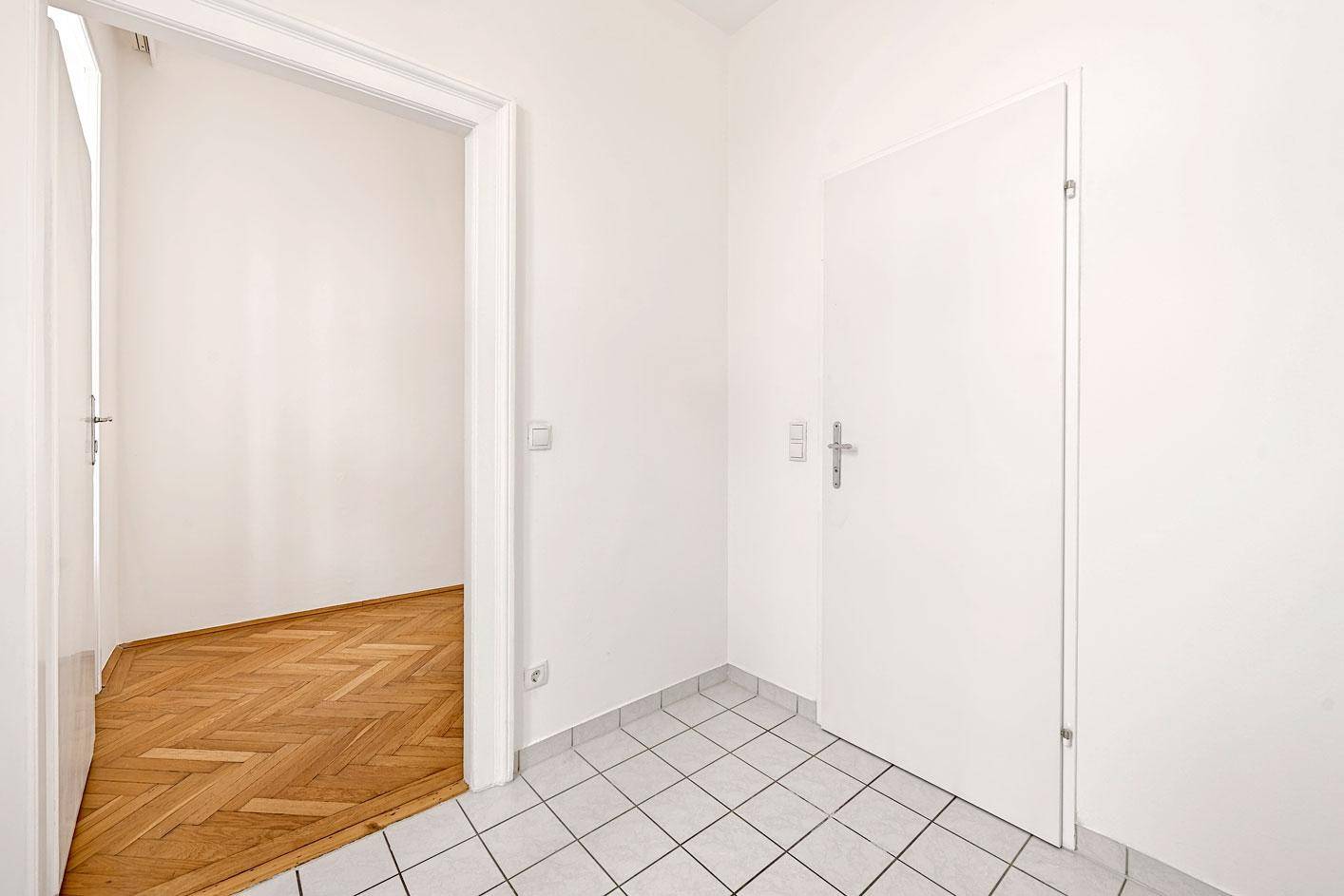 Eingangsbereich mit weißen Fliesen und einer Tür, die zu einem Zimmer mit Parkettboden führt.
