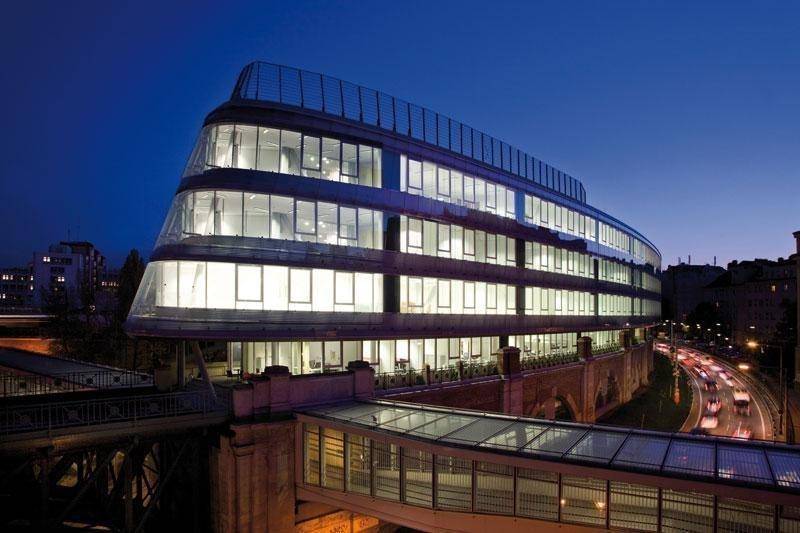 Beleuchtetes Bürogebäude bei Nacht mit geschwungener Architektur und Stadtlichtern im Hintergrund, beeindruckende Ansicht.