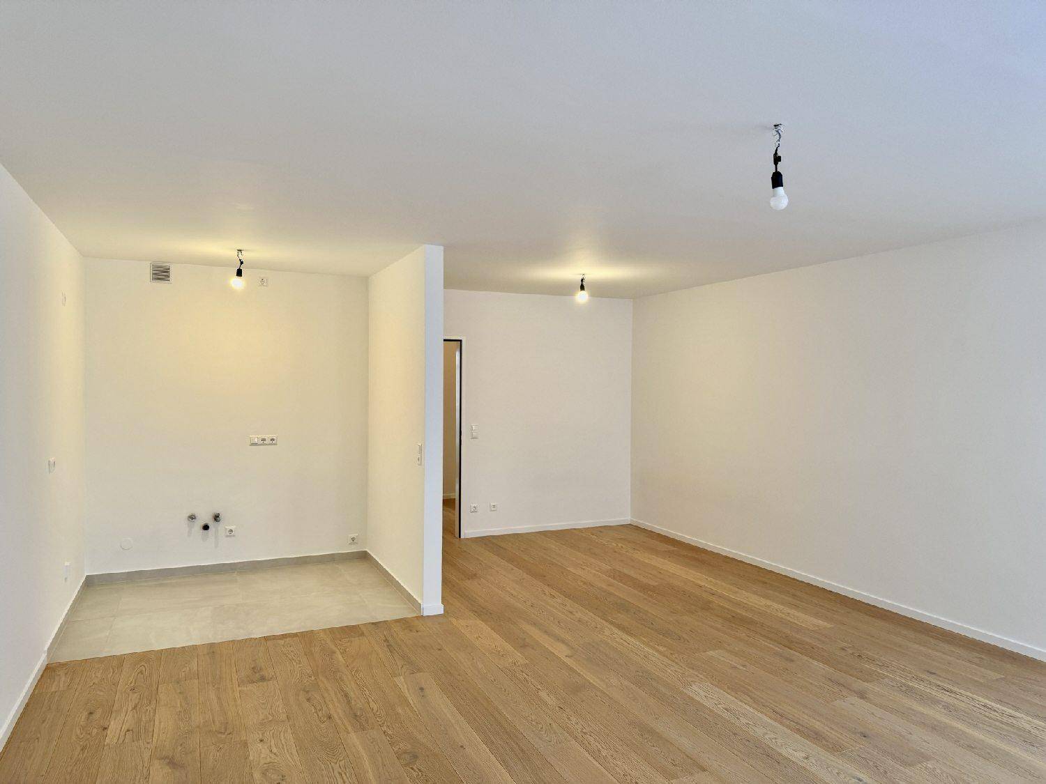 Heller Raum mit Parkettboden und vorbereitetem Bereich für eine moderne Küche.
