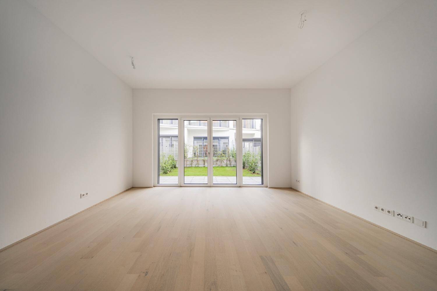 Geräumiges, leeres Zimmer mit großen Fenstern und Blick auf den grünen Gartenbereich.