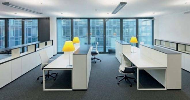 Helles Großraumbüro mit weißen Schreibtischen, gelben Lampen und großen Fensterfronten, die viel Tageslicht hereinlassen.