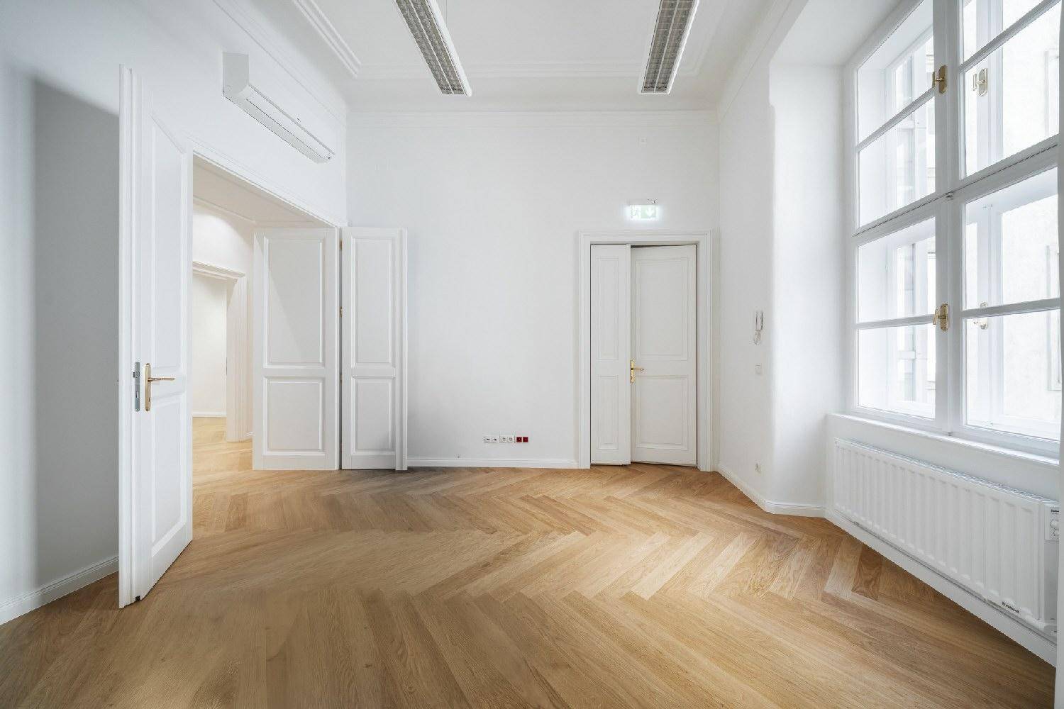 Heller, leerer Raum mit Fischgrätparkett, weißen Wänden, Klimaanlage und großem Fenster.