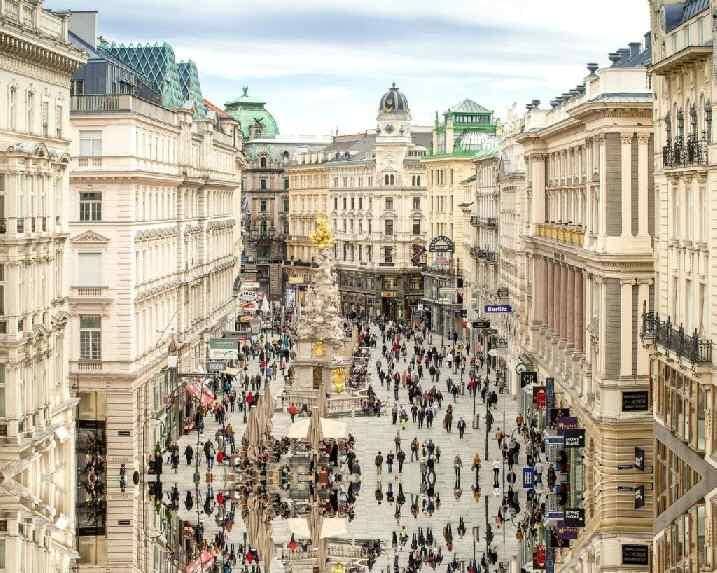 Belebte Einkaufsstraße in einer historischen Stadt mit prächtigen Gebäuden und einem zentralen Denkmal.