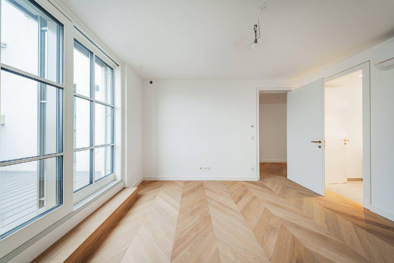 Modernes Zimmer mit Fischgrätparkett, großen Fenstern und Zugang zu weiteren hellen Räumen.