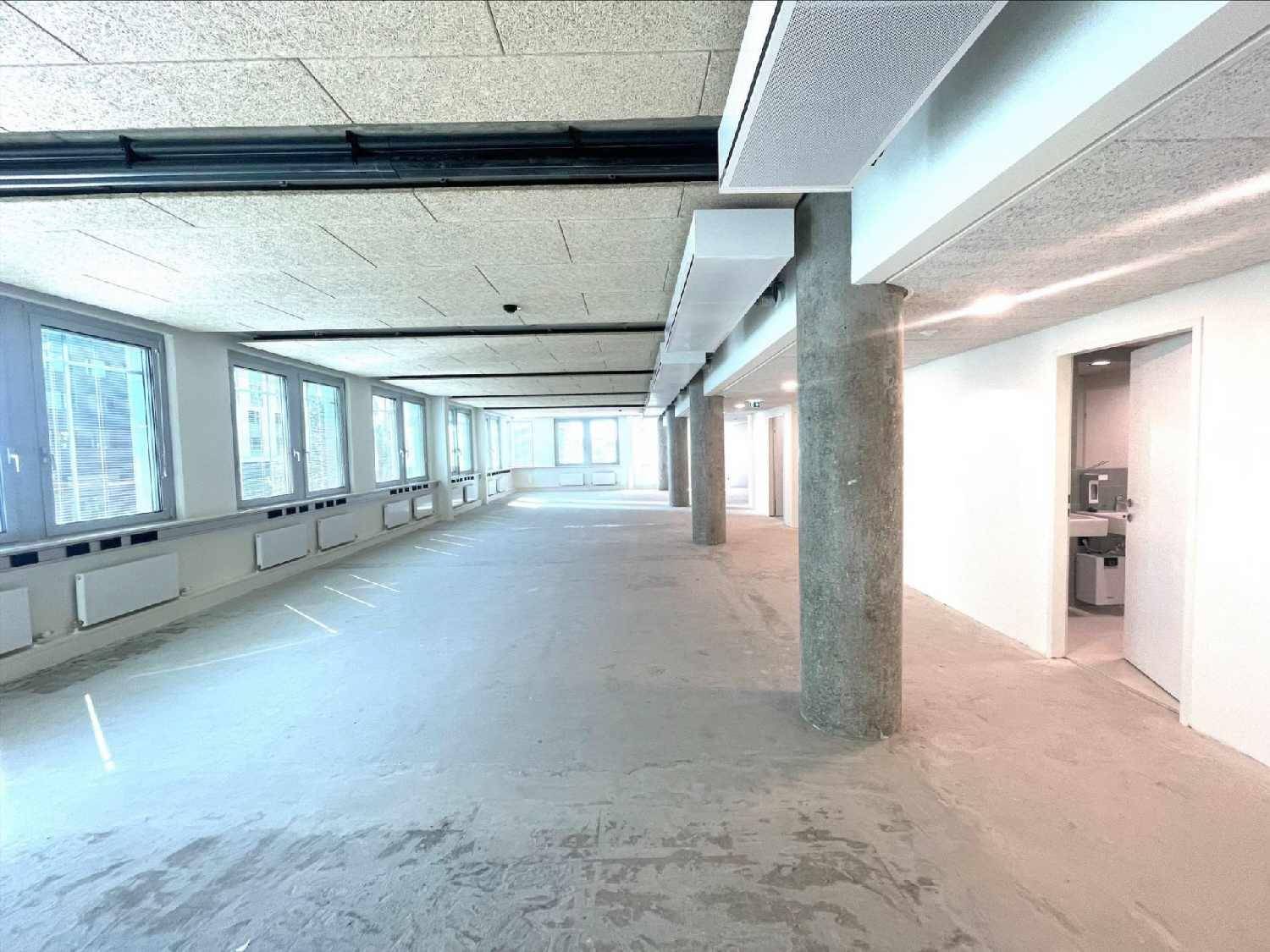 Heller Büroraum mit Betonboden und Akustikdecke, bietet Zugang zu einem Nebenraum mit Waschbecken.