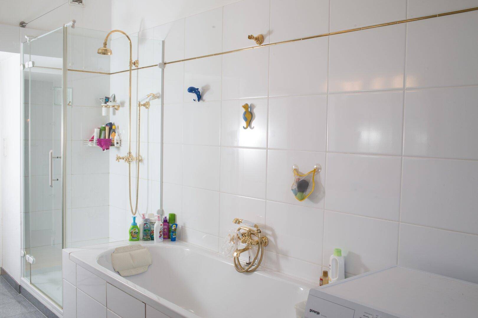 Badezimmer mit weißen Fliesen, Badewanne, Duschkabine und goldenen Armaturen, inklusive Waschmaschine.
