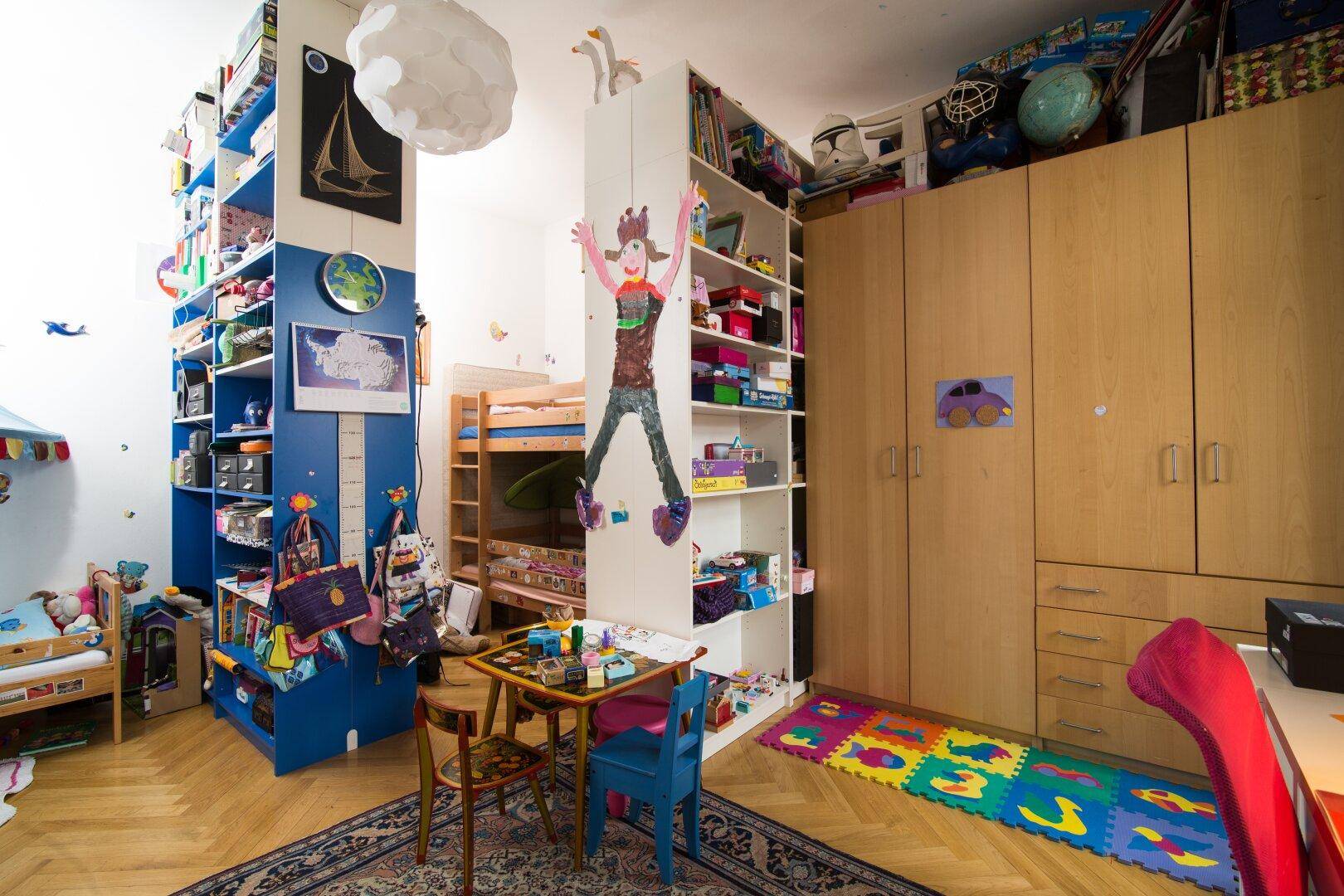 Farbenfrohes Kinderzimmer mit Hochbett, Spielbereich und vielen Regalen für Bücher und Spielzeug.