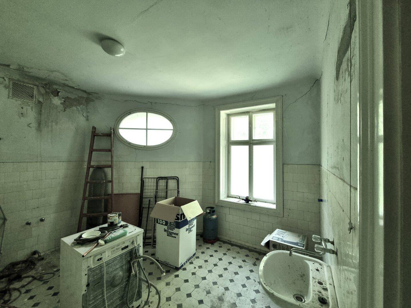 Badezimmer in renovierungsbedürftigem Zustand mit Fliesenboden und zwei Fenstern.