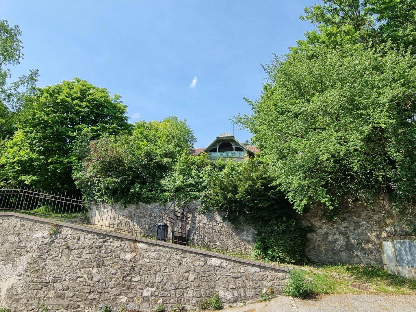 Historisches Gebäude hinter einer Steinmauer und Bäumen, mit Blick auf die Umgebung.