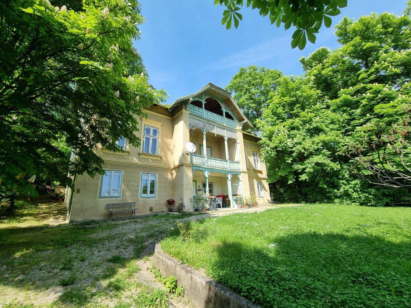 Historisches Gebäude mit gelber Fassade, umgeben von üppigem Grün und Bäumen unter blauem Himmel.