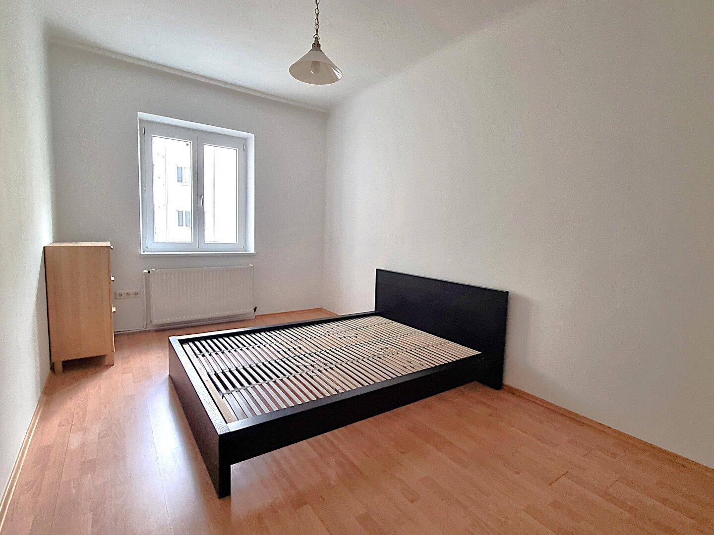 Schlafzimmer mit Bettrahmen, Kommode und Fenster, bietet eine einfache und funktionale Einrichtung.