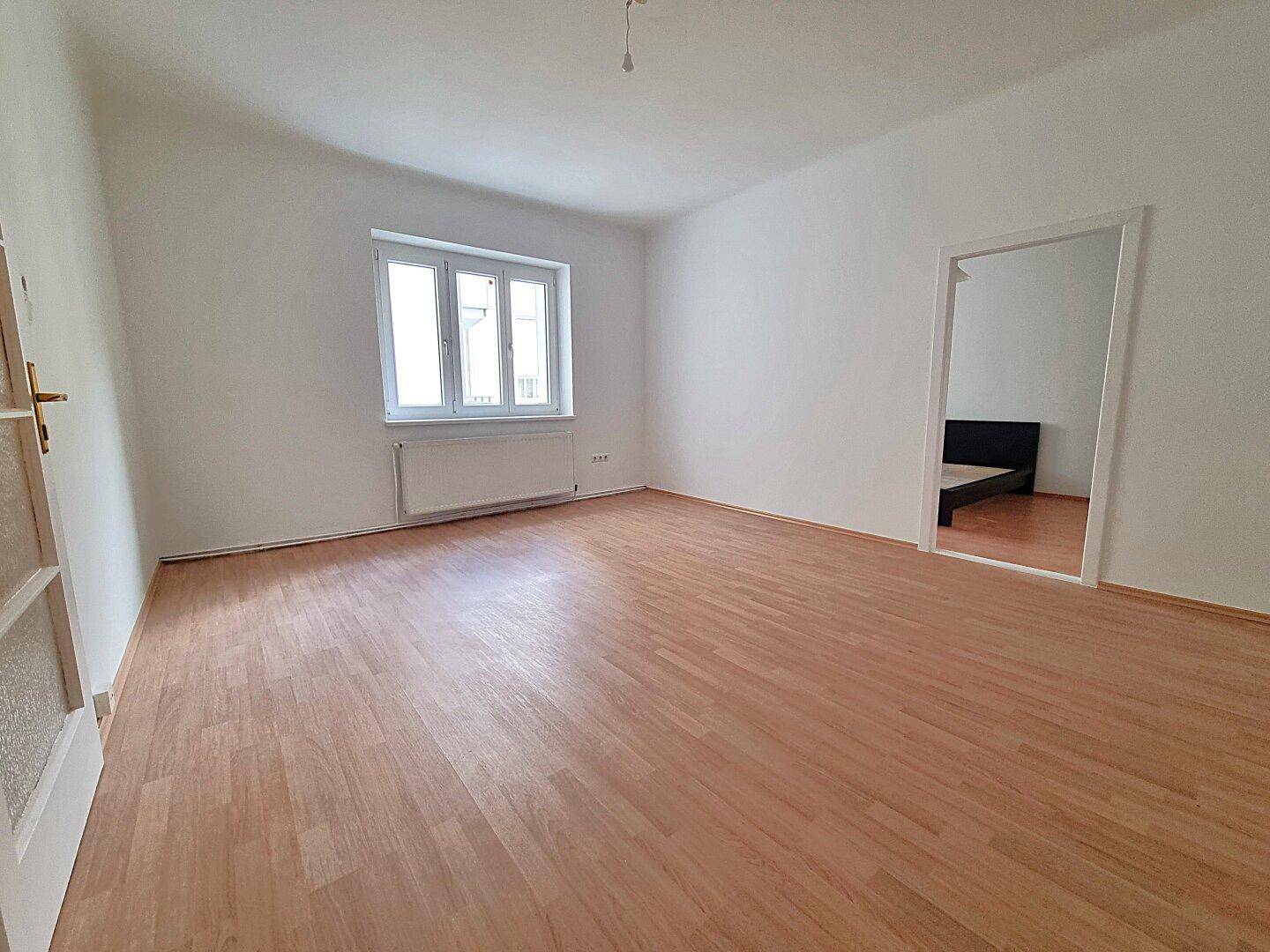 Heller Wohnraum mit Laminatboden und offenem Zugang zu einem angrenzenden Schlafzimmer.
