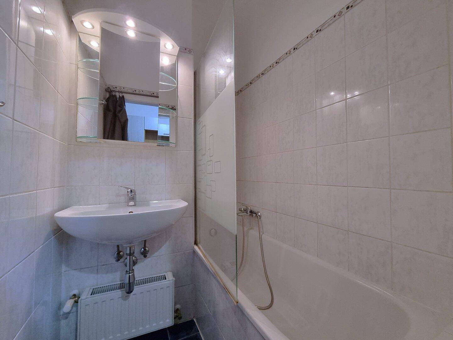 Badezimmer mit weißen Fliesen, Waschbecken, Badewanne mit Duschwand und Spiegelschrank.