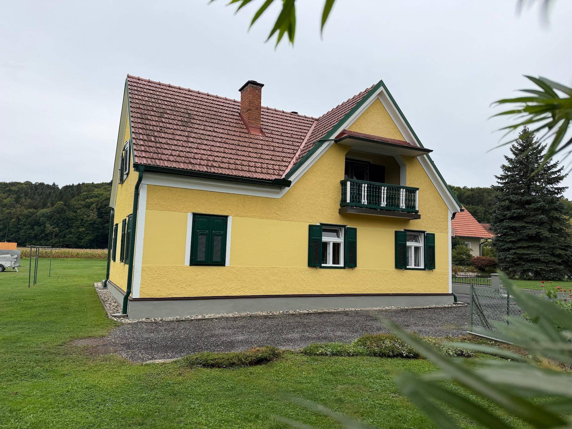 Seitenansicht des gelben Hauses mit rotem Ziegeldach, grünen Fensterläden und einem kleinen Balkon.