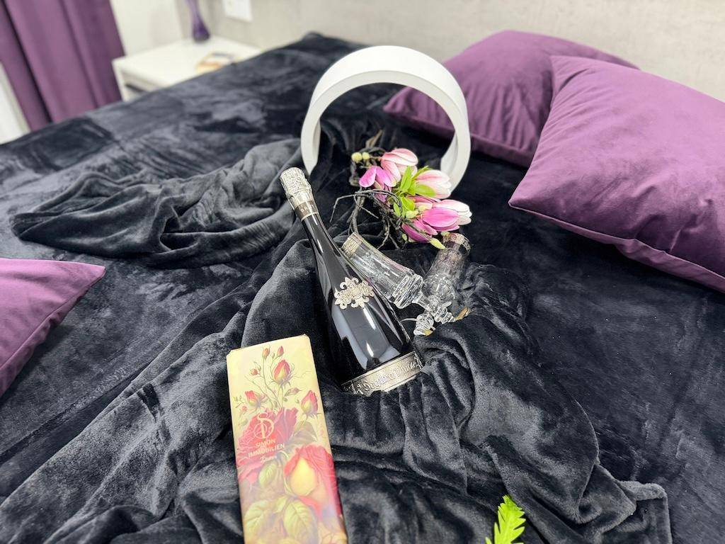 Nahaufnahme einer Champagnerflasche mit Gläsern und Blumen auf dem dunklen Bett im Schlafzimmer.