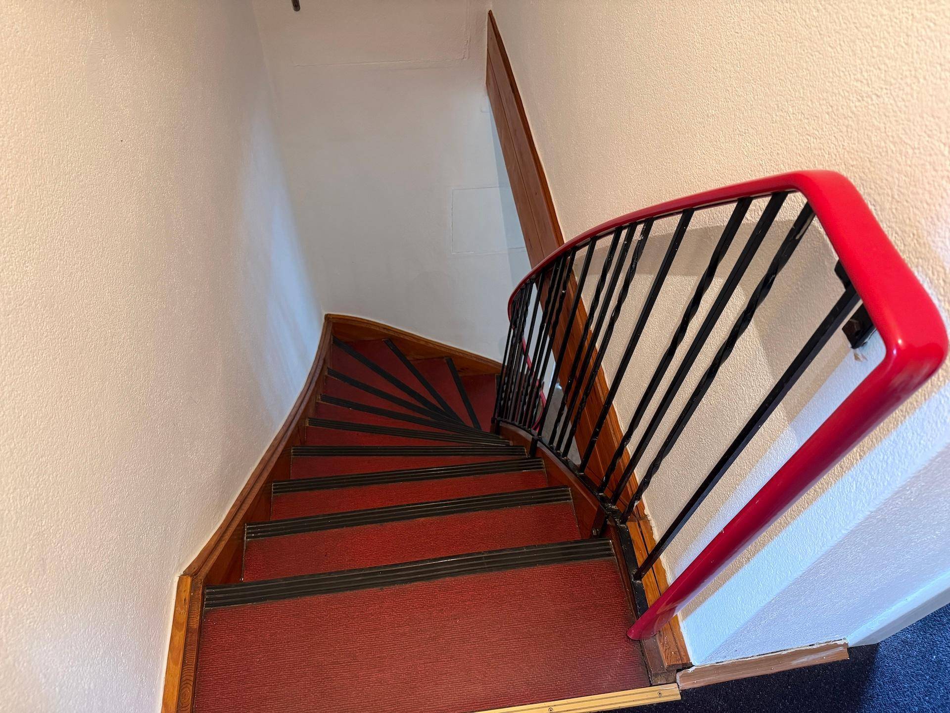Holztreppe mit rotem Teppich und einem markanten rot-schwarzen Geländer führt in die obere Etage.