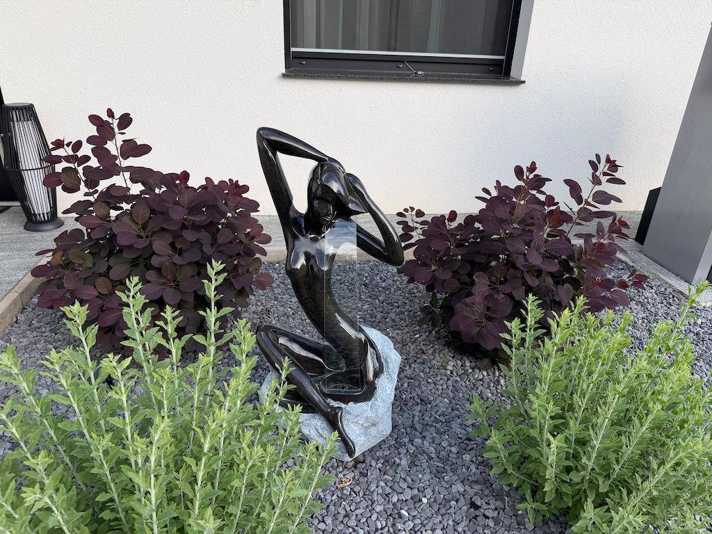 Detailansicht einer modernen Gartendekoration mit Skulptur, umgeben von Zierpflanzen und Kies.