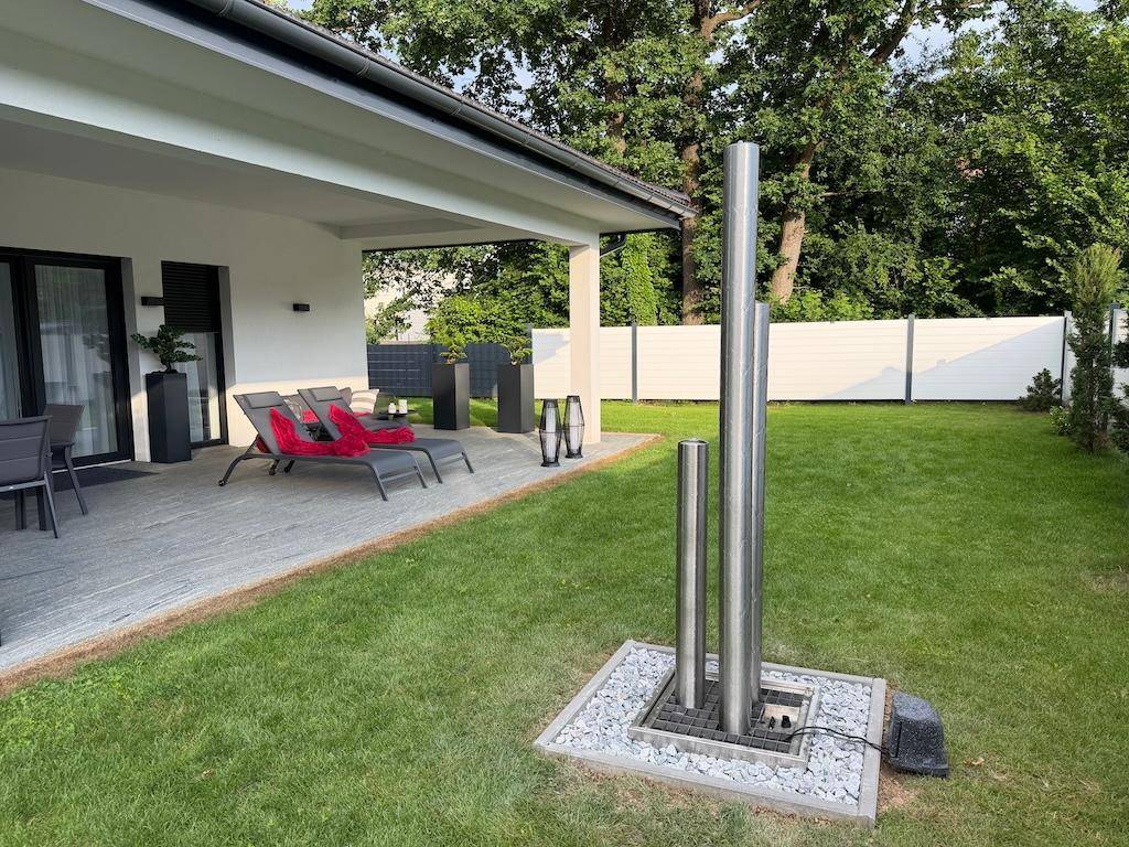 Sonniger Gartenbereich mit überdachter Terrasse, Liegestühlen und einem modernen Wasserspiel.