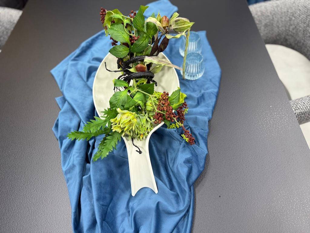 Elegante Tischdekoration mit grünen Pflanzen und blauer Stoffbahn auf einem dunklen Esstisch.