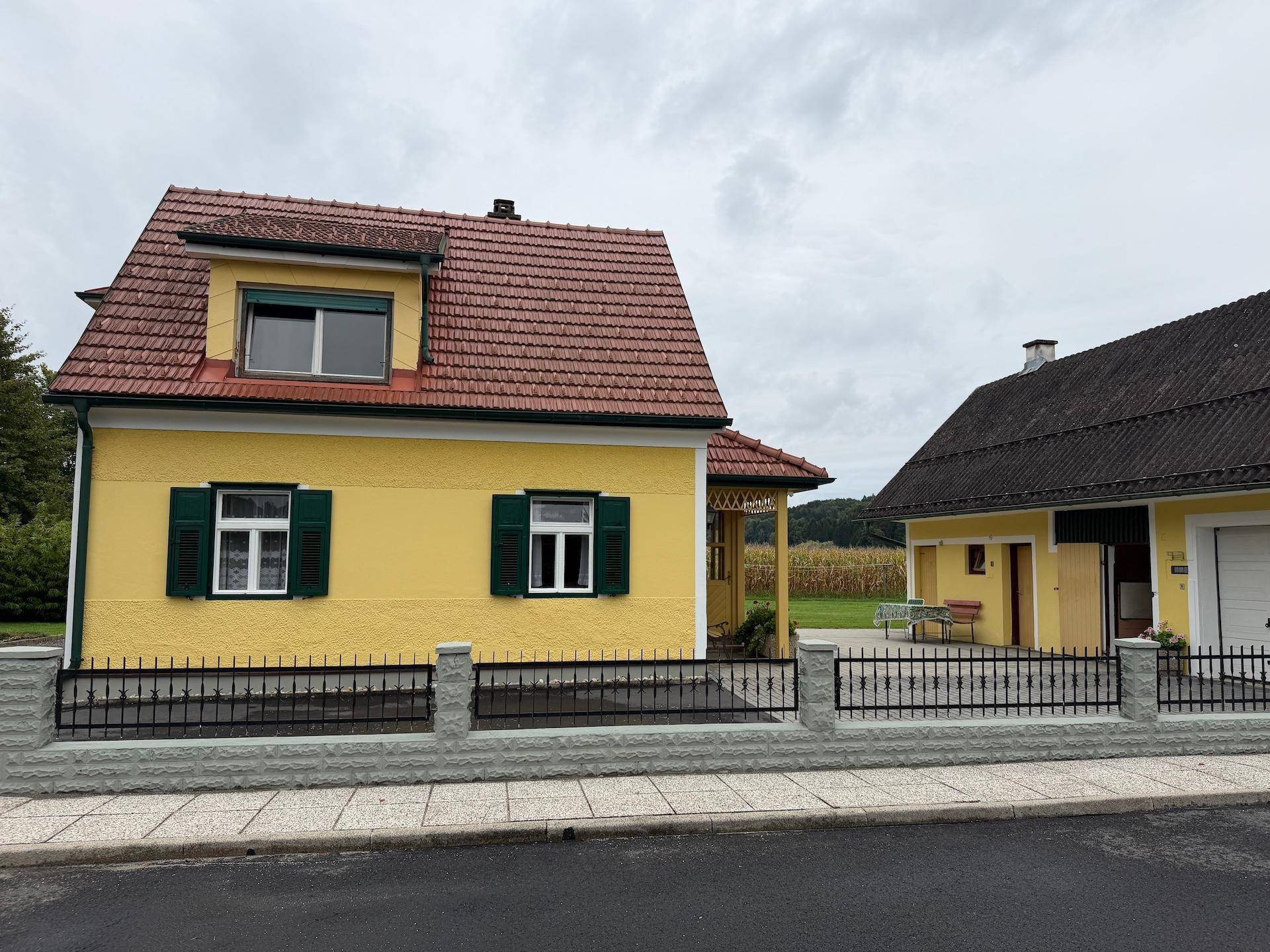 Vorderansicht des Hauses mit gelber Fassade, rotem Dach und einem Zaun zur Straße hin.