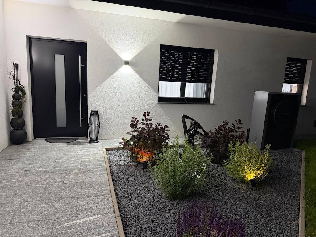 Beleuchteter Eingangsbereich des Hauses bei Nacht mit moderner Haustür und dekorativer Bepflanzung.