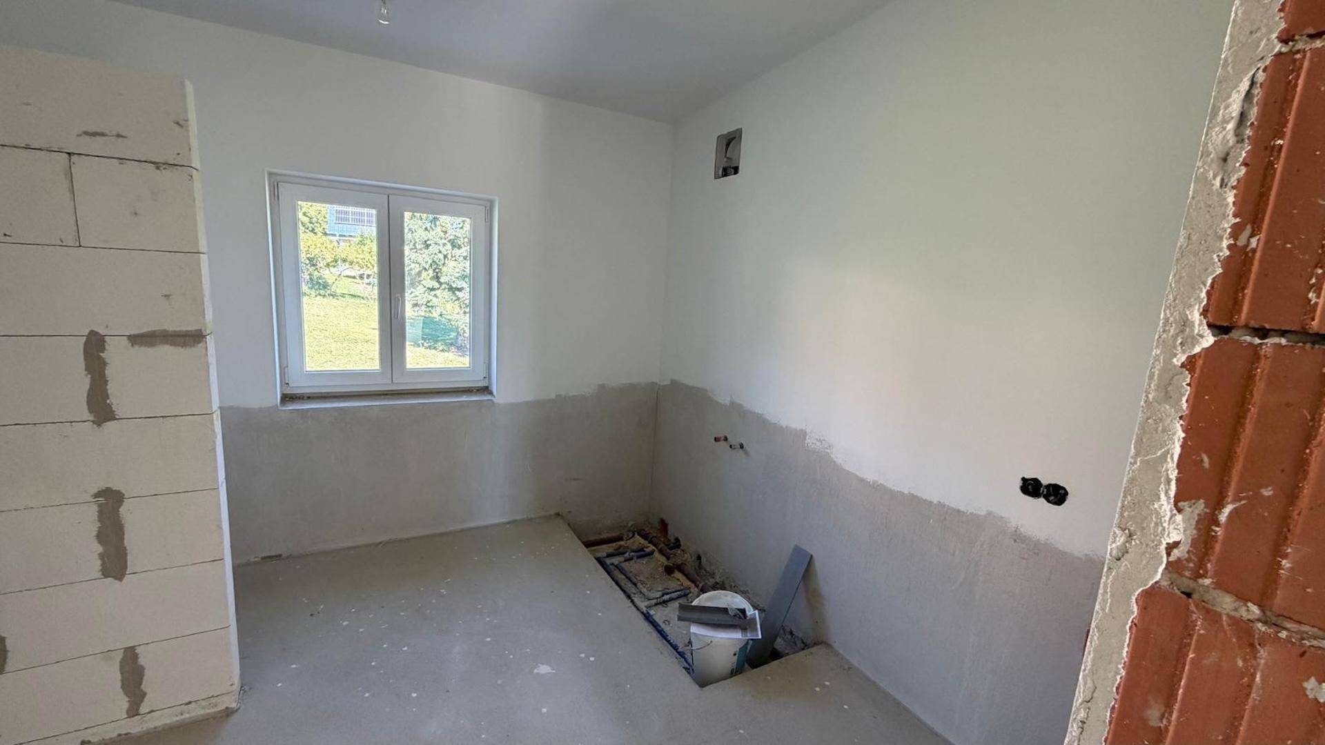 Unfertiges Badezimmer mit sichtbaren Sanitäranschlüssen und einem Fenster für natürliche Belüftung.