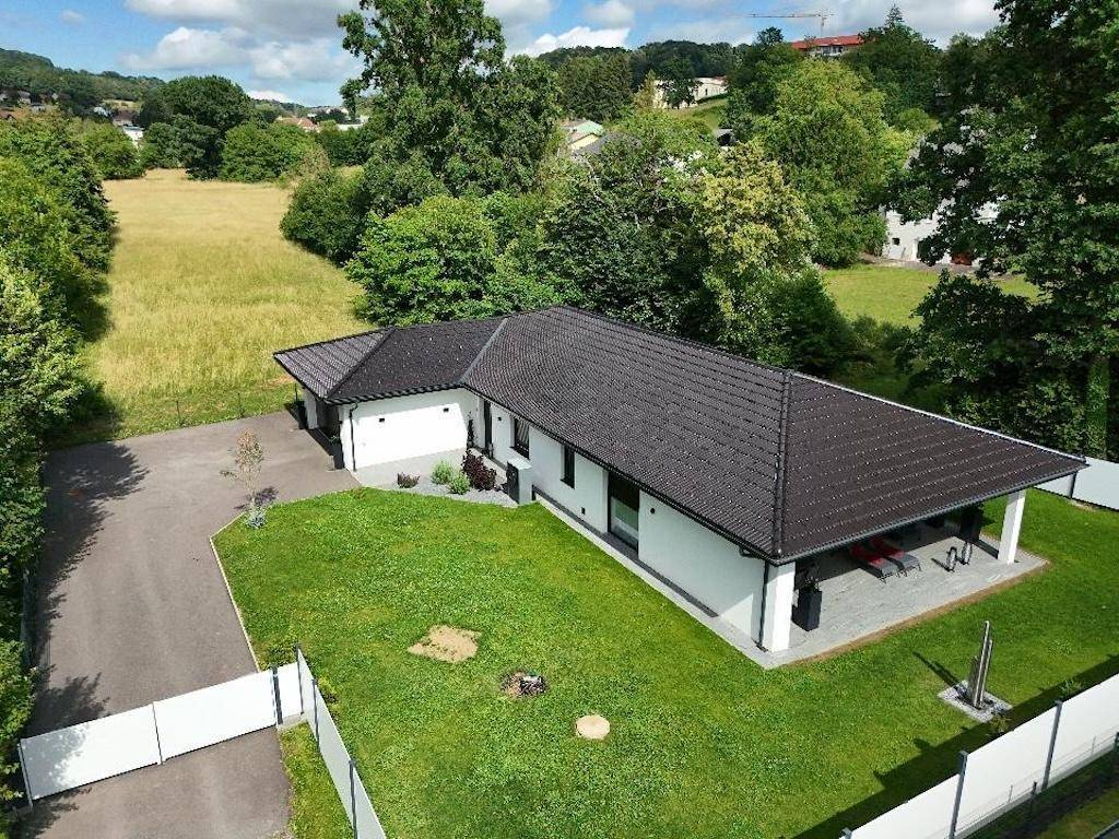 Luftaufnahme eines modernen Einfamilienhauses mit großem Garten und Zufahrt, eingebettet in grüne Landschaft.