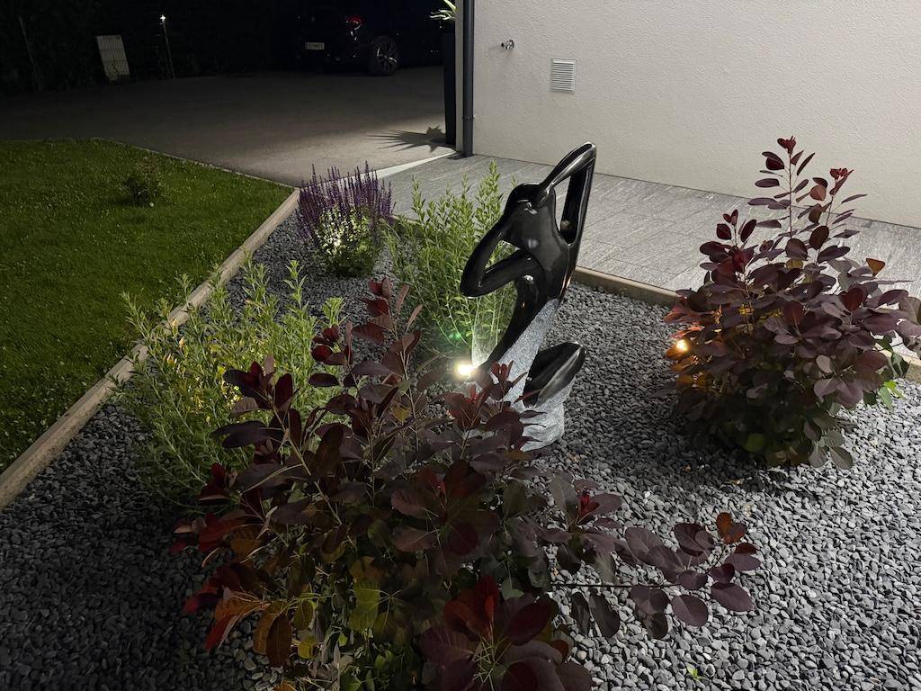 Beleuchtetes Blumenbeet mit Zierkies und einer modernen Skulptur im Vorgartenbereich.