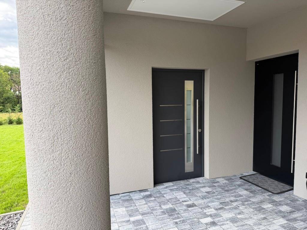 Moderner Hauseingang mit grauer Tür, Glaseinsätzen und gepflastertem Vorplatz unter dem Carport.