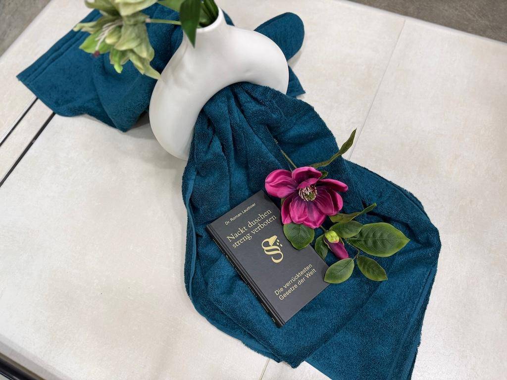 Detailaufnahme von tealfarbenen Handtüchern, einer Blume und einem Buch auf hellem Fliesenboden.