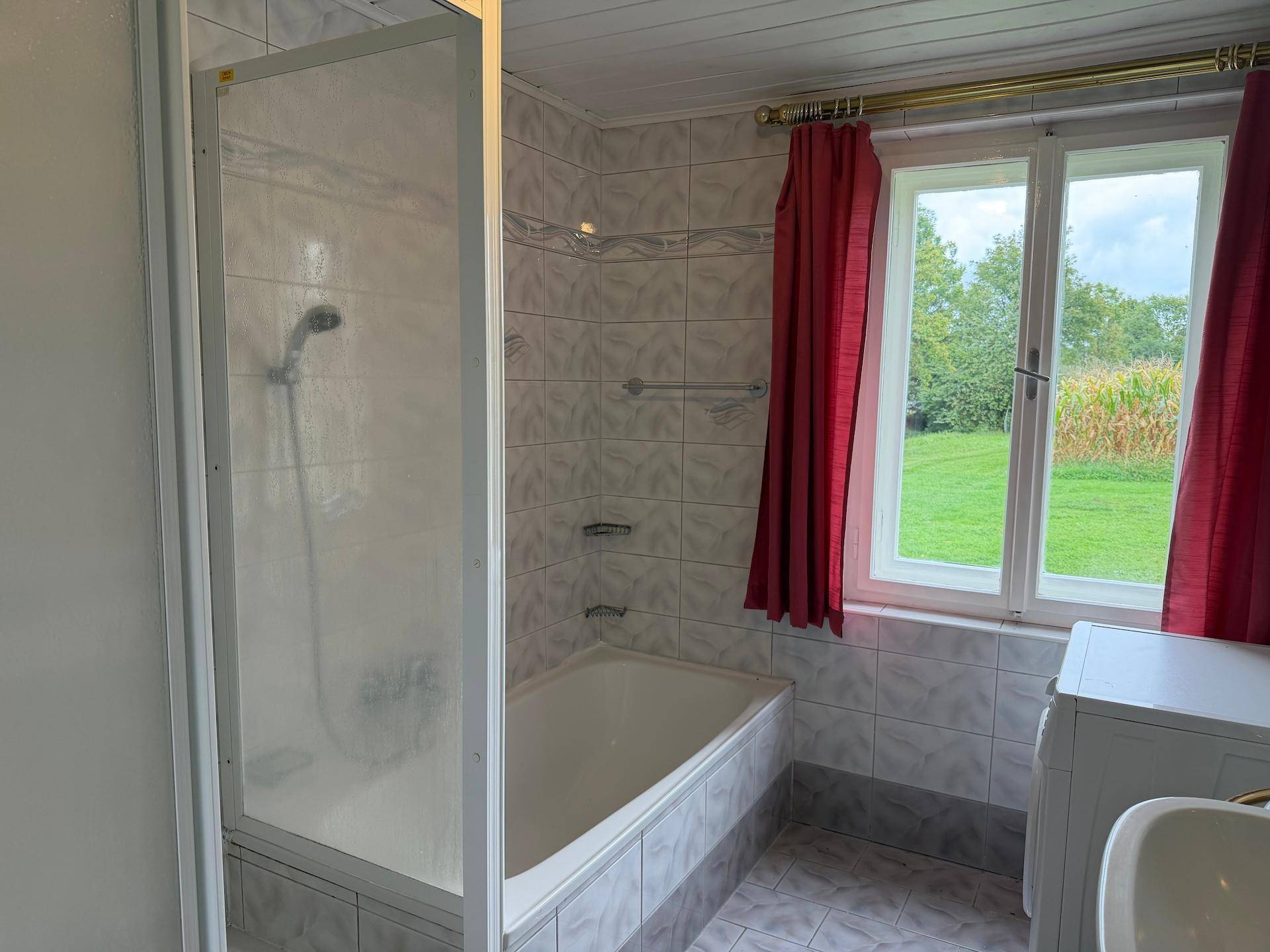 Badezimmer mit Badewanne und Duschabtrennung, umgeben von Fliesen und einem Fenster mit Blick ins Grüne.