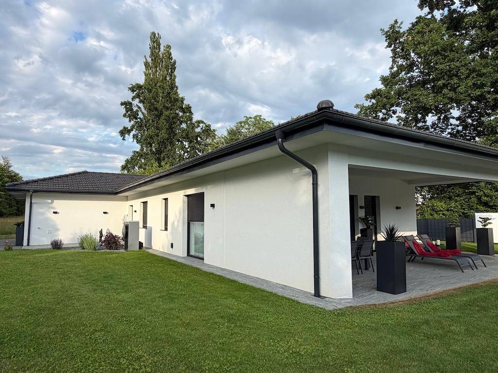 Modernes Einfamilienhaus mit weitläufigem Rasen und großer überdachter Terrasse, umgeben von viel Grün.