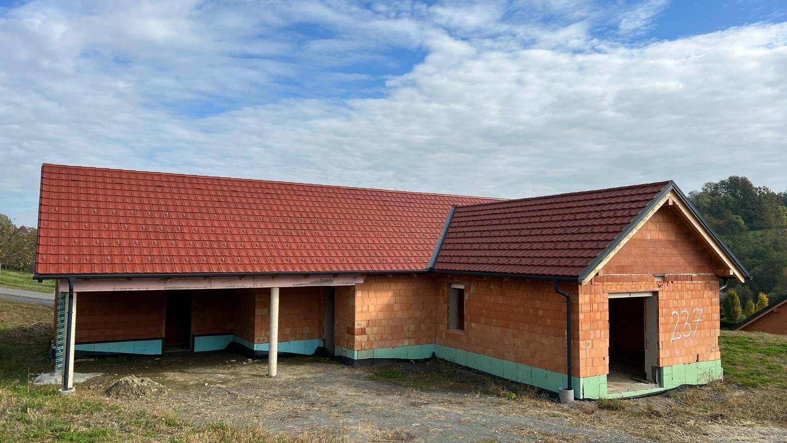 Seitenansicht des Rohbaus mit integriertem Carport und rotem Ziegeldach in ländlicher Umgebung.