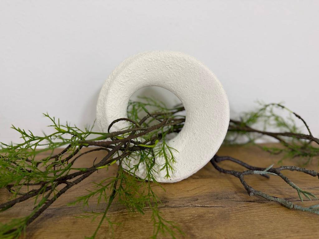 Detailaufnahme einer weißen, ringförmigen Dekoration mit grünen Zweigen auf einer Holzoberfläche.