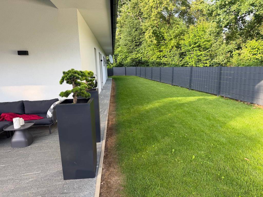 Langer Rasenbereich neben der überdachten Terrasse, umgeben von einem modernen Zaun und Pflanzen.