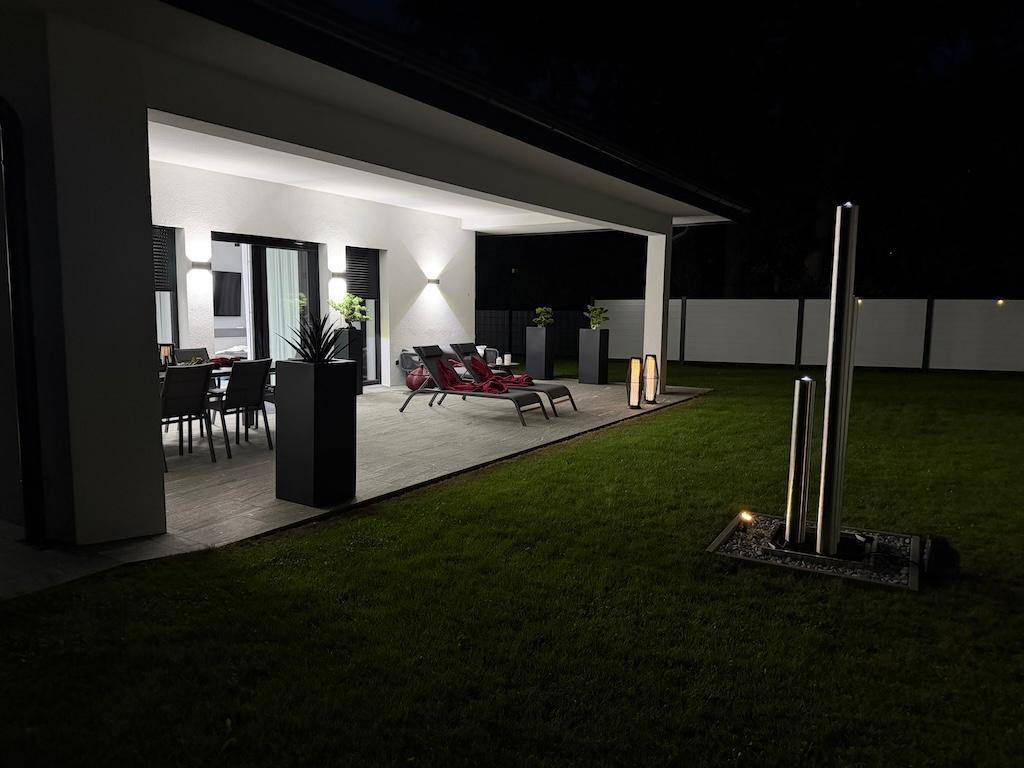 Beleuchtete Terrasse am Abend mit Essbereich, Liegestühlen und dekorativen Pflanzen.