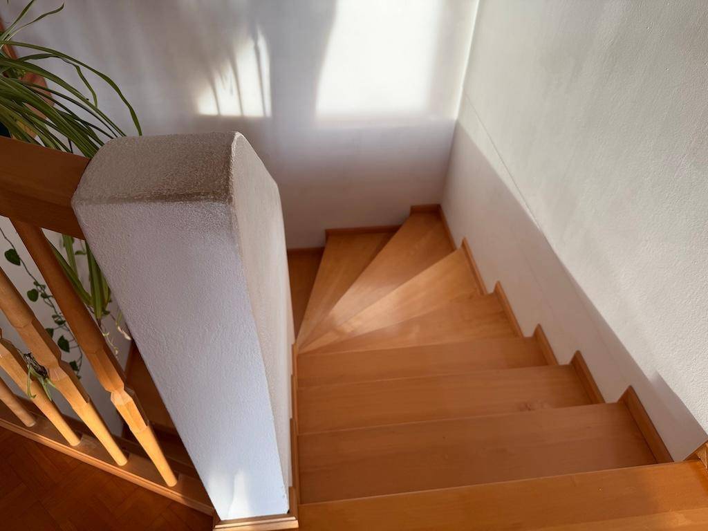Holztreppe führt nach unten, mit weißer Wand und Holzgeländer, gut beleuchtet.