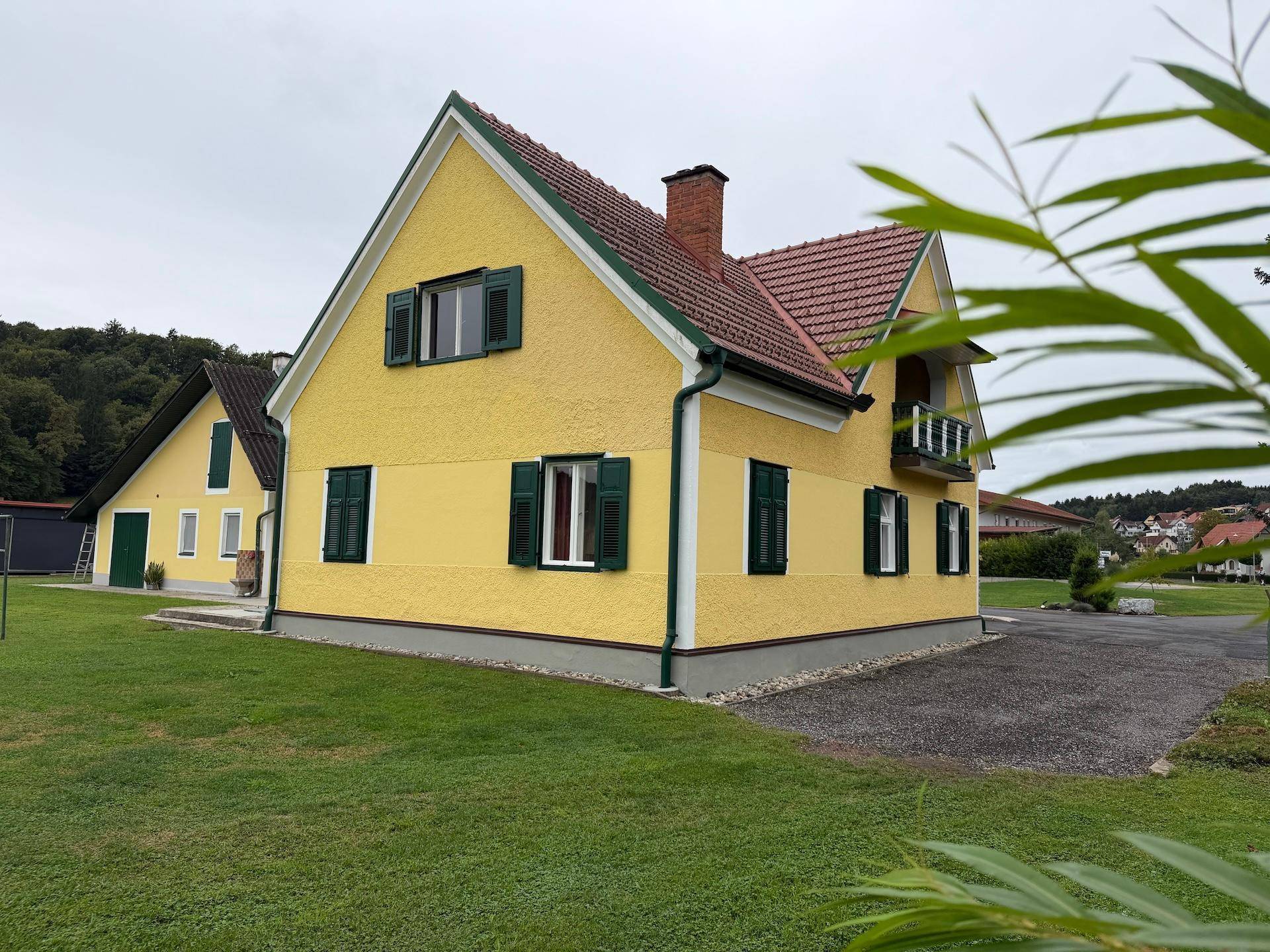 Seitenansicht des gelben Hauses mit roten Dachziegeln, grünen Fensterläden und einem kleinen Balkon.