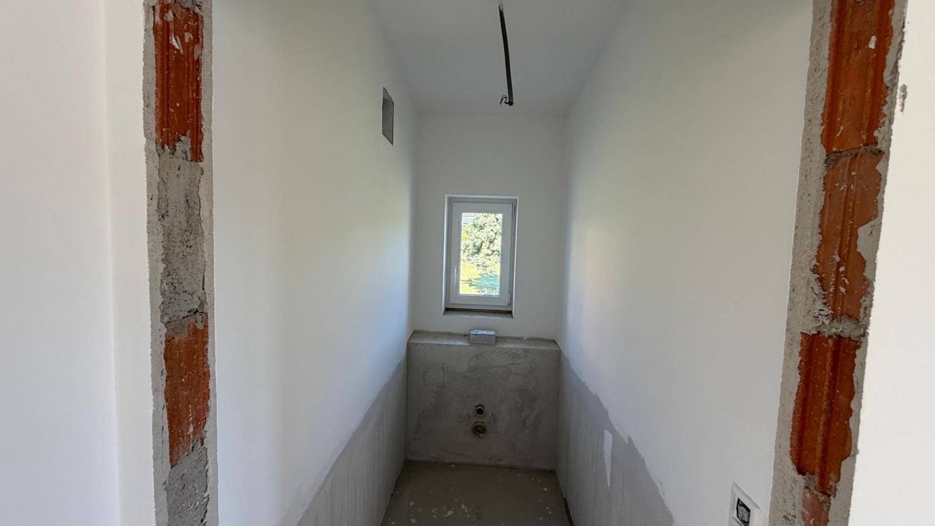 Schmaler Raum im Rohbau, wahrscheinlich ein Gäste-WC, mit Fenster und Sanitäranschlüssen.