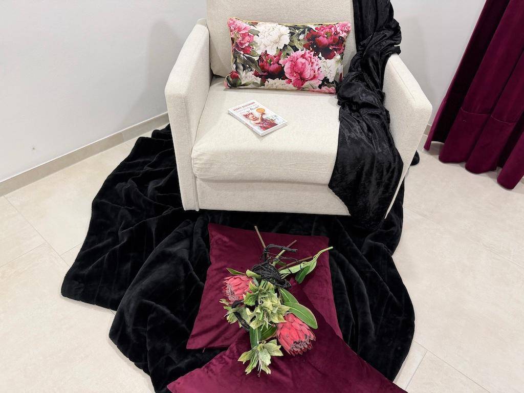 Gemütlicher Sessel mit floralem Kissen und dunkler Decke, umgeben von weichen Textilien.