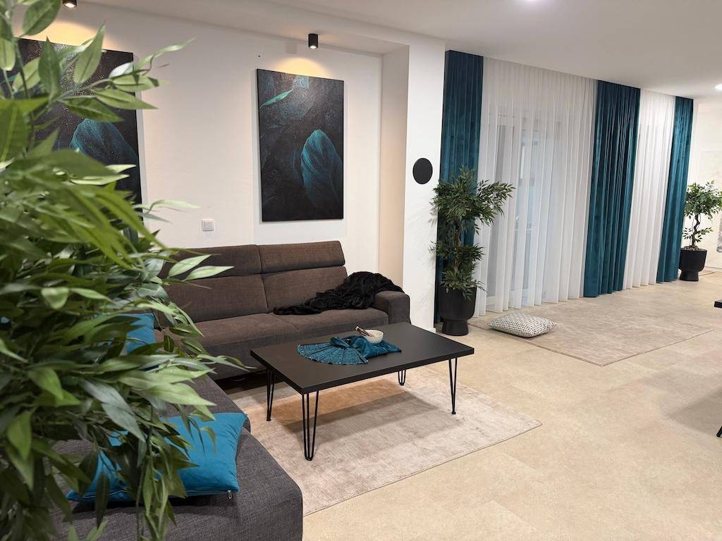 Weitläufiges Wohnzimmer mit beigem Sofa, grünen Pflanzen und großen Fenstern mit Vorhängen.