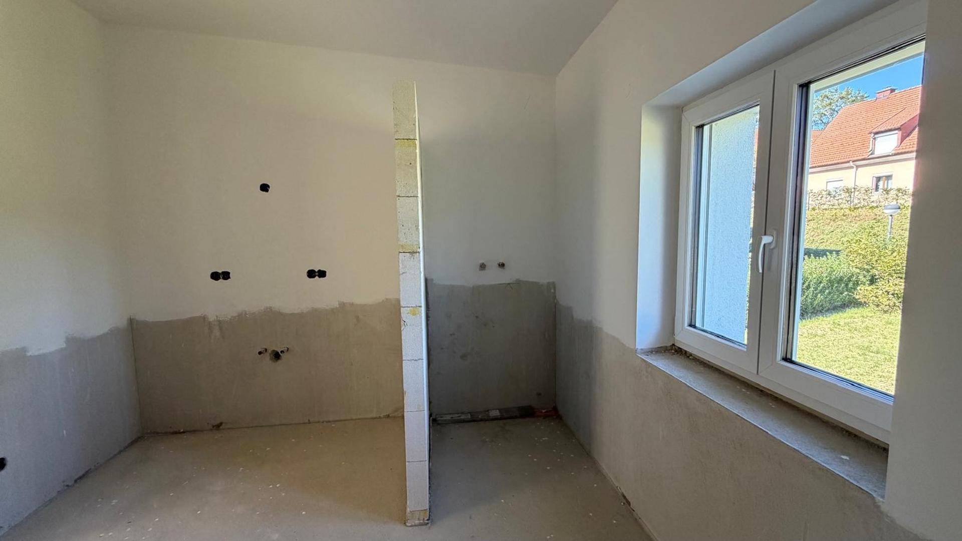 Unfertiges Badezimmer mit vorbereiteten Anschlüssen und einem Fenster, bereit für die Installation.