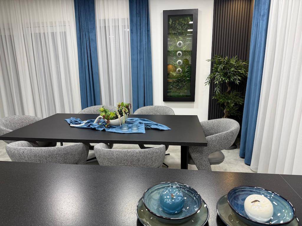 Essbereich mit dunklem Tisch, grauen Stühlen und einer dekorativen Pflanzenwand, umgeben von Vorhängen.