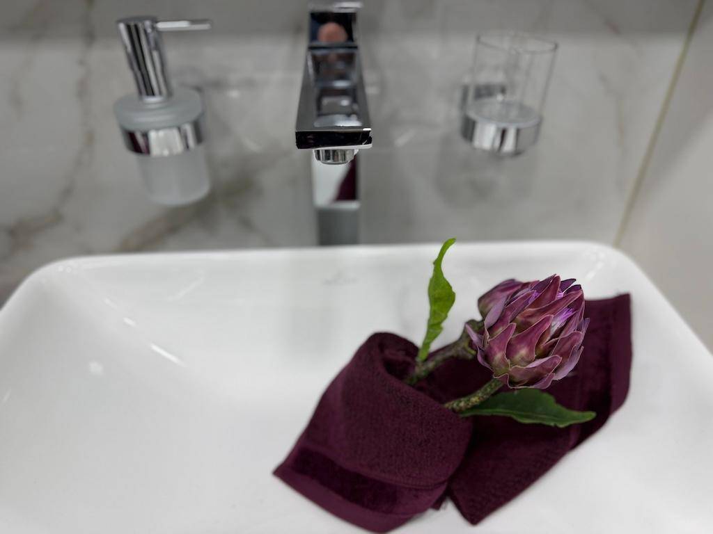 Nahaufnahme eines modernen Waschbeckens mit Armatur und einer dekorativen Blume auf einem lila Handtuch.