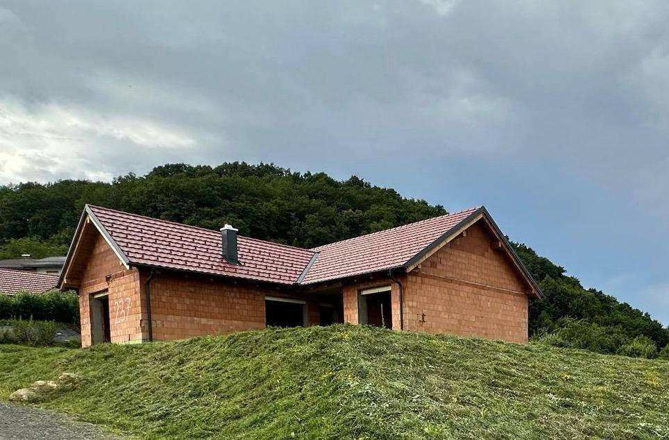 Rohbau eines Einfamilienhauses auf einem Hügel mit bewaldetem Hintergrund unter bewölktem Himmel.