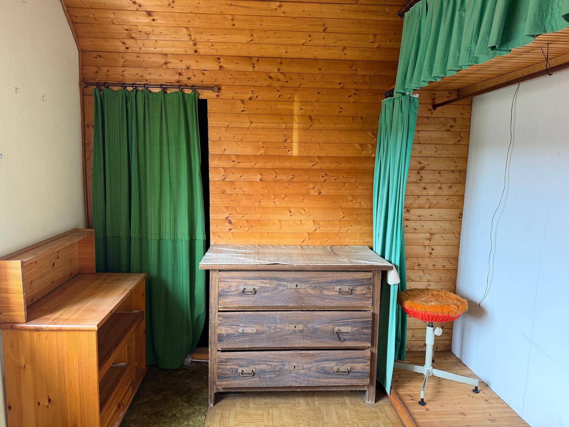 Rustikaler Raum mit Holzvertäfelung, alter Kommode und grünem Vorhang, möglicherweise ein Schlafzimmer.