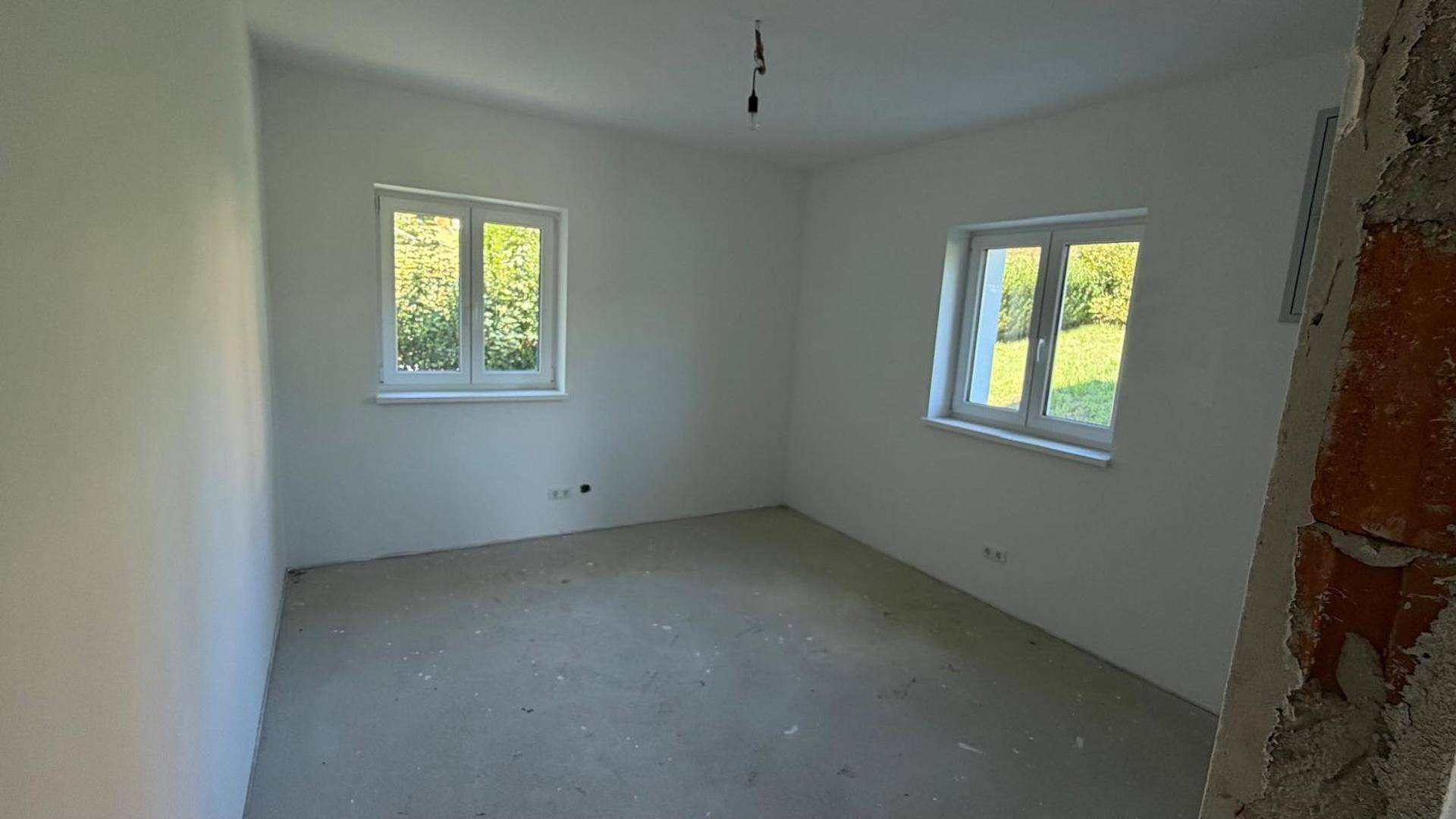 Helles, unfertiges Zimmer mit zwei Fenstern und weiß gestrichenen Wänden.